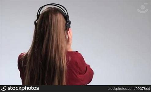 Half-length of a young charming brunette woman with beautiful long hair listening to music with massive headphones on her head, rear view. The stunning girl is turning back and smiling happily at the camera while enjoying the track on the radio.