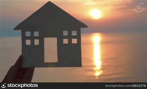 Hand holding white cut paper house against the sea at sunset. Unrecognizable.