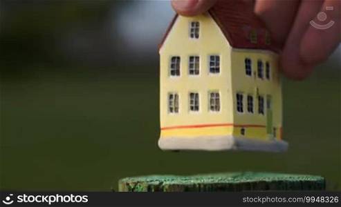 Hand putting a toy house on small stub on blurry natural background. Real estate, country house or mortgage symbol
