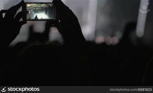 Hands in the crowd holding smartphone and shooting musical performance. Bright illumination on the stage