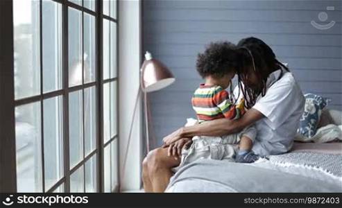Handsome African man sitting on the bed in the bedroom and talking with his toddler son while the sweet boy is sitting on the father's lap. Happy stylish daddy with dreadlocks talking with his little kid, head to head, and smiling in a domestic interior background.