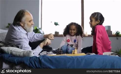 Handsome grandpa with beard offering cute mixed race granddaughter a possibility to choose between black or white chess pieces, hiding them in his hands. Grandfather and little girls getting ready to play chess while sitting on bed at home. Dolly.