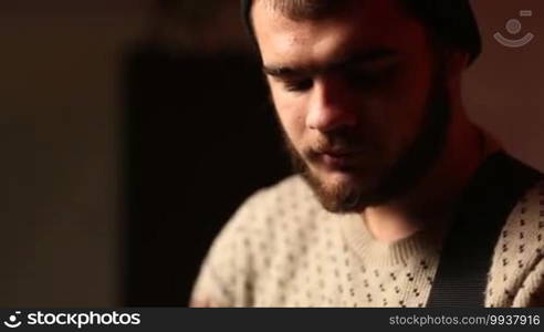 Handsome man with beard and hat playing guitar in garage. Young guitarist rehearsing with the band and practicing playing acoustic guitar. Close up hipster male hands playing musical instrument.