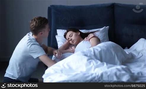 Handsome teenager sitting beside the bed and gently waking his pretty girlfriend, stroking her hair with love and tenderness. Hipster man waking his sleeping woman in the morning.