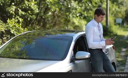 Handsome young businessman in necktie talking on smartphone while leaning on parked car over rural landscape background. Successful entrepreneur in formal wear communicating with business partner using mobile phone outdoors during business road trip.