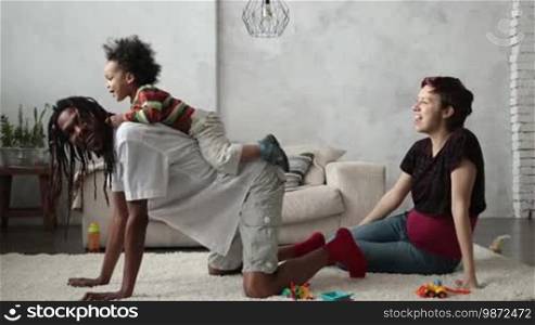 Happy African American father with dreadlocks giving cute mixed race toddler son piggyback ride in the living room while Caucasian pregnant mother watching them playing. Multiethnic family spending time together at home. Side view. Slow motion.