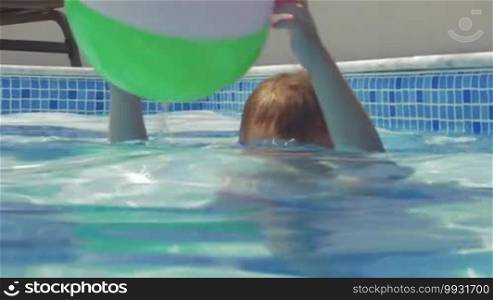 Happy and active little boy in goggles playing with wind ball in outdoor swimming pool on hot sunny day