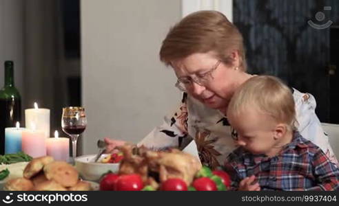 Happy charming senior woman feeding her grandson on Thanksgiving. Loving grandmother sitting at festive table with her cute toddler grandson on her knees and feeding child with healthy food.