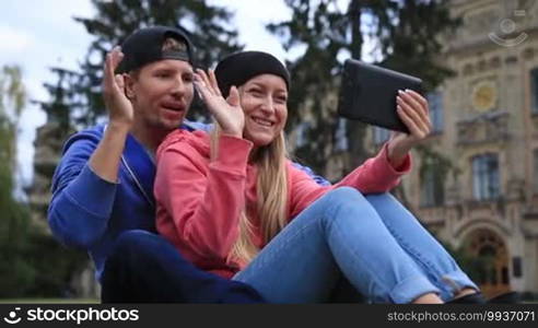 Happy couple of students sitting on the grass in the park outside university and communicating through video chat with their friends online using electronic digital tablet. Smiling students greeting their online teacher and waving hello with hands.