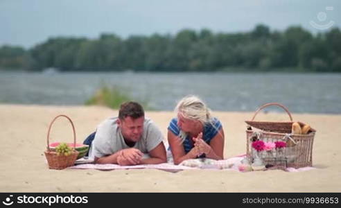 Happy family on vacation lying on a blanket on the beach and chatting during a picnic. Son comes near his parents and kisses them with tenderness