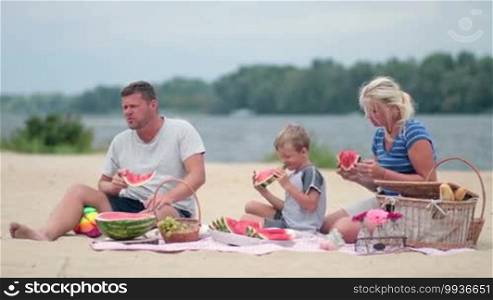 Happy family sitting on a colorful blanket on the beach during summer vacation and having a picnic. Family with a child eating watermelon.
