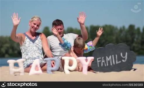 Happy family with son on summer vacation sitting on the beach and waving hello with hands over blue sky background. Foreground word happy made of fabric padded letters and word family written with chalk on black board