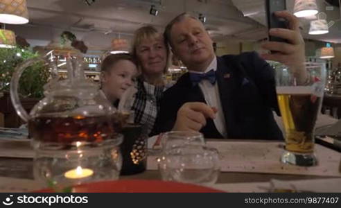 Happy grandma, grandpa, and grandson taking a family selfie with a cellphone during a celebratory dinner in the restaurant