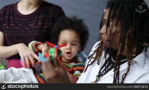 Happy interracial family with parents and curly little toddler son playing with colorful toys in the bedroom at home. Smiling stylish African dad with dreadlocks and mixed race cute child having fun together indoors closeup.