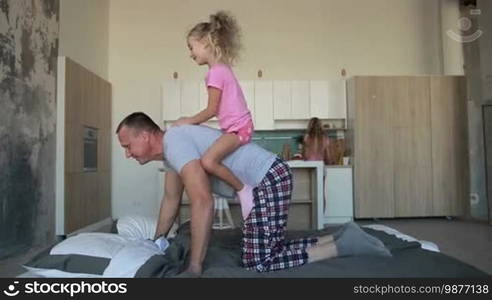 Happy mad ride on father's back. Excited daughter riding on father's back at home. Playful father in pajamas piggybacking joyful little girl while playing with child on bed in the morning. Beautiful mother preparing breakfast on background. Slo mo.