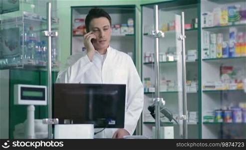 Happy male pharmacist standing behind counter, communicating on phone with client and browsing on computer in pharmacy. Cheerful pharmacy worker talking to customer by smartphone and typing on computer keyboard.