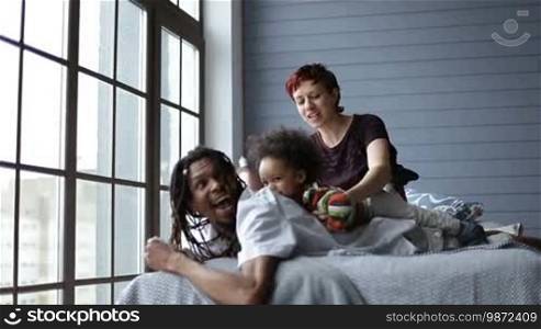 Happy mixed family relaxing in the bedroom at home. Smiling African father with dreadlocks lying on bed, Caucasian mother putting their mixed-race toddler son on daddy's back and tickling her boy. Joyful family with child lounging at home interior.