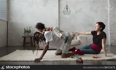 Happy mixed race toddler son riding on father's back in the modern apartment. Playful African American daddy with dreadlocks and joyful kid playing piggyback at home. Multiethnic family lounging and having fun together. Slow motion.