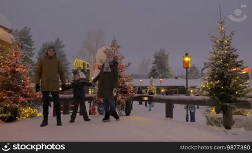 Happy parents with child in snowy park decorated with Christmas lights. They holding hands, smiling and looking to the camera