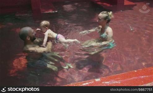 Happy people, family, father, mother, child, baby in swimming pool. Mom and dad helping daughter, little girl, infant swim. Fun, leisure, recreation, lifestyle, parenthood.
