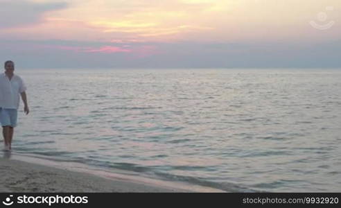 Happy senior man walking barefoot in the shore water at sunset. Loving couple holding hands and enjoying summer time