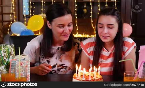 Happy teen girl and mother with birthday cake at anniversary party. Lighting candles on birthday cake. People celebrating birthday concept. Slow motion hand held movement
