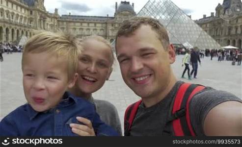 Happy tourists family making selfie video near the The Louvre museum in Paris. Parents smiling and son waving hand to say hello