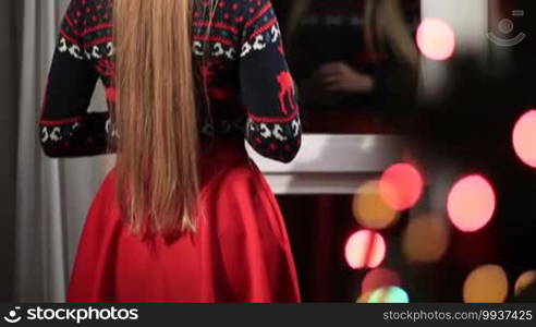 Happy woman standing in living room and waving from window for welcoming return of her beloved husband home on Christmas Eve. Back View. Blurred colorful bokeh circles of Christmas lights on tree.