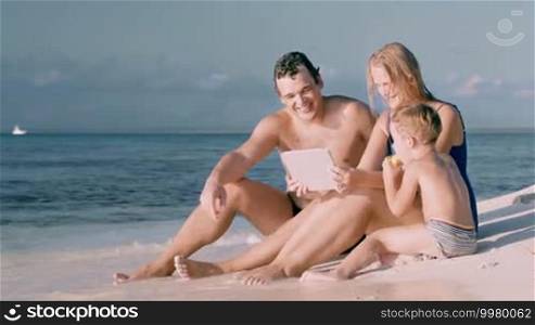 Happy young parents and little son sitting on the beach and watching video or photos on tablet computer. Water washing their feet