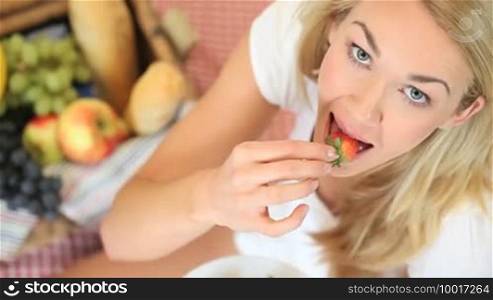 High angle view of a beautiful blonde woman sitting on a rug at a picnic enjoying eating a strawberry