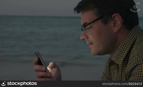 Hipster style man in glasses and check shirt using smart phone on the beach in the evening. He texting sms or chatting in social network service. Sea surf in background