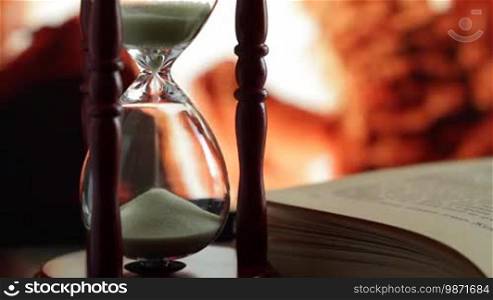 Hourglass and book on the background of burning fireplace.
