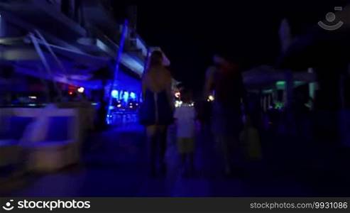 Hyperlapse shot of parents and little son holding hands and walking along vibrant night street, passing by outdoor cafes and stores