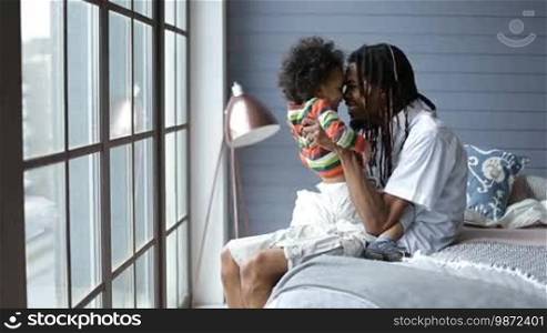 Joyful African American father with dreadlocks sitting on the bed and holding his toddler son on laps while playing with boy. Happy man fooling around with adorable mixed race child and rubbing nose together at home interior.