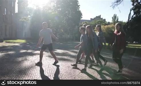 Joyful classmates going through university campus to study. Smiling group of students entering university building. Attractive friends walking through the park on their way to college. Slow motion. Steadicam stabilized shot.