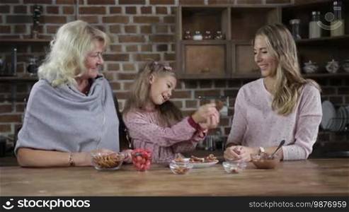 Joyful daughter giving homemade chocolate cookies to beautiful mother and smiling grandmother in the kitchen. Cheerful little girl sharing delicious cookies with her loving mom and grandma while multi-generation family spending leisure at home.