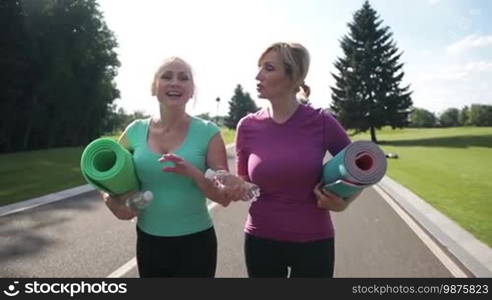 Joyful fitness adult women with exercise mats and bottles of water going to make sport and fitness outdoors. Attractive senior females talking while walking through the park to exercise in the park. Slow motion. Steadicam stabilized shot.