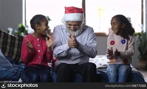 Joyful grandfather in Santa hat with beard giving Christmas gift boxes to his excited mixed-race granddaughters at home. Two happy little sisters embracing their affectionate granddad on the bed as family celebrating Christmas in domestic room.