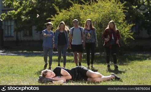 Joyful group of college students waking up their friend while handsome male hipster is sleeping on green grass on university campus. Smiling university friends with backpacks and books waking up their tired classmate, who fell asleep on park lawn
