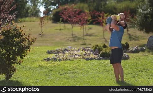 Joyful hipster father in casual clothes throwing his infant boy high up in the air over beautiful landscape background. Happy dad and toddler baby son lifting him up, playing and having fun in summer park while enjoying leisure together in sunlight.