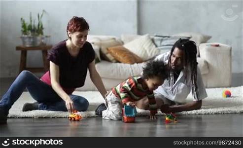 Joyful interracial family with cute mixed race toddler son spending leisure together at home. Happy pregnant Caucasian mother, African American dad and little sweet child sitting on the floor in the living room and playing together with colorful toys.