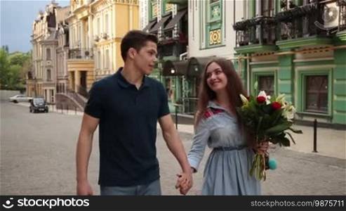Joyful young hipster couple spending leisure together. Charming woman with bouquet of flowers holding hand of her handsome man walking along city street during romantic date. Slow motion. Steadicam stabilized shot.