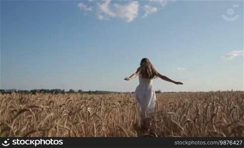 Joyful young woman in white dress with amazing long blond hair running through wheat field in summer against amazing skyline background. Positive beautiful female having fun and enjoying beauty of nature in countryside. Slow motion. Stabilized shot.