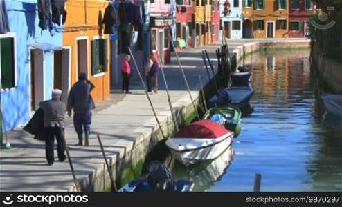 Kanal in Burano, Fußgänger laufen am Hafen entlang.