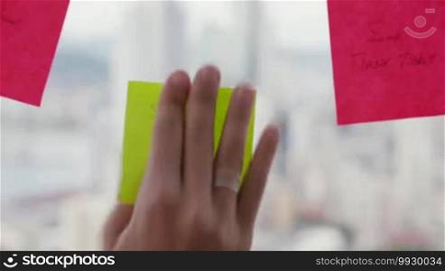 Latin American woman working as a secretary in a modern office with a beautiful view of the city, sticking adhesive notes with tasks on a skyscraper window. Rack focus from the city to her hand. Close-up shot