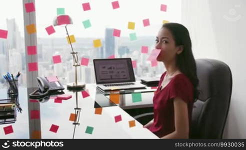 Latin American woman working as a secretary in a modern office with a beautiful view of the city, sticking adhesive notes with tasks on a skyscraper window. The girl feels stressed, holding a note with a sad emoticon on her mouth. Medium shot