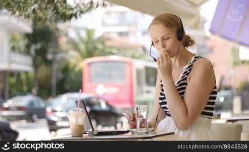 Laughing attractive blond woman wearing a headset sitting with a laptop at an open-air restaurant enjoying refreshments and talking to her friend using Skype