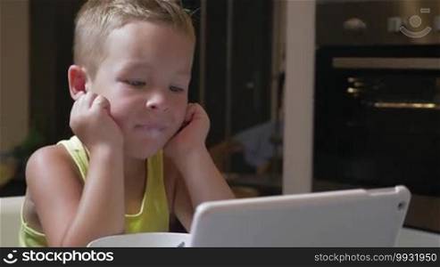 Little blond boy having a meal in the kitchen. He is watching cartoons on a touchpad to entertain himself while eating