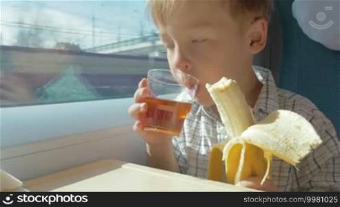 Little blond boy sitting near the window eats banana and drinks juice while traveling by train, then he talks