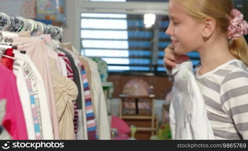 Little blond girl shopping for clothes in a clothing store, looking at a sweater. Side view